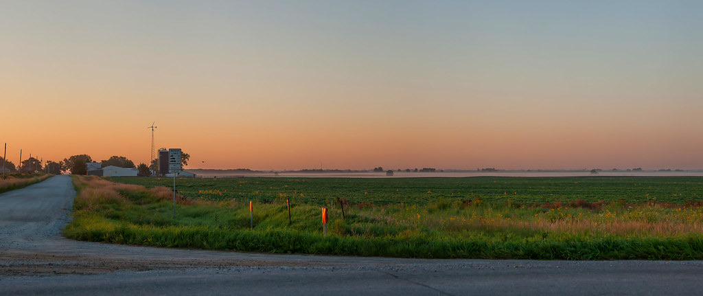 A Missouri farm field at first light, warm orange sky over a quiet rural road at dawn
